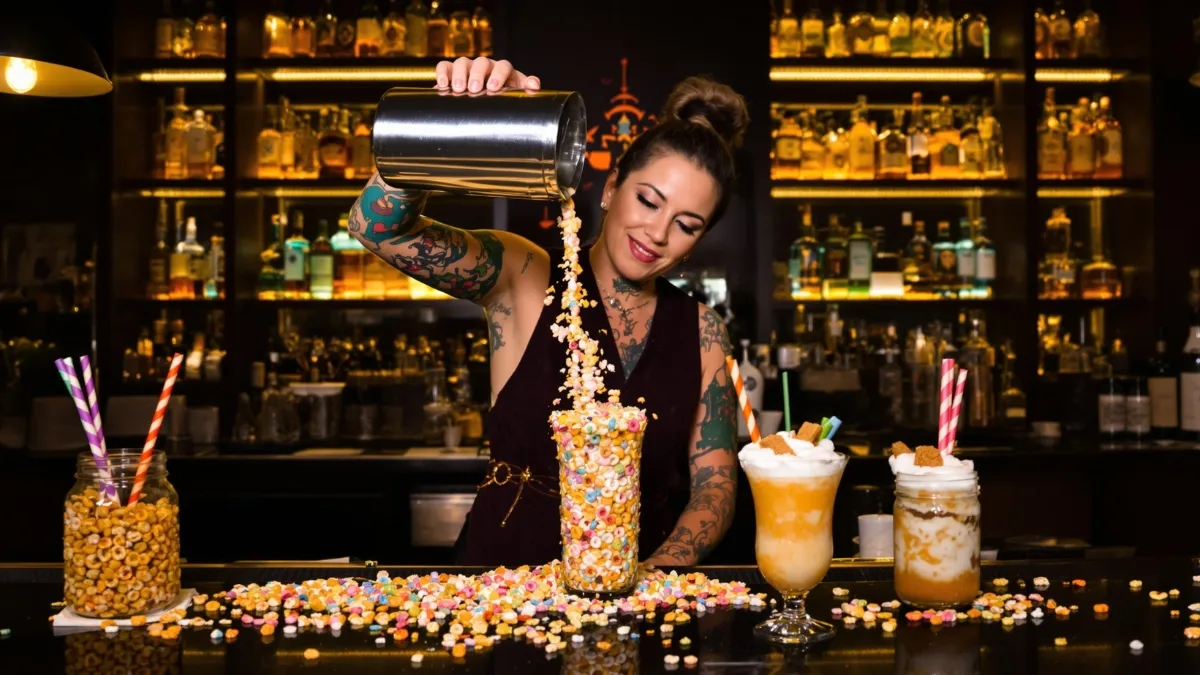 A stylish mixologist in her early 30s, with vibrant sleeve tattoos and a vintage-inspired